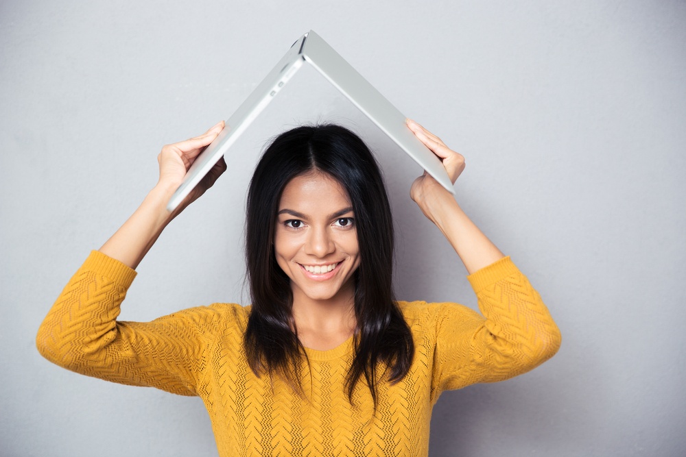 Happy woman in sweater holding laptop above her head like a roof over gray background. Looking at camera.jpeg